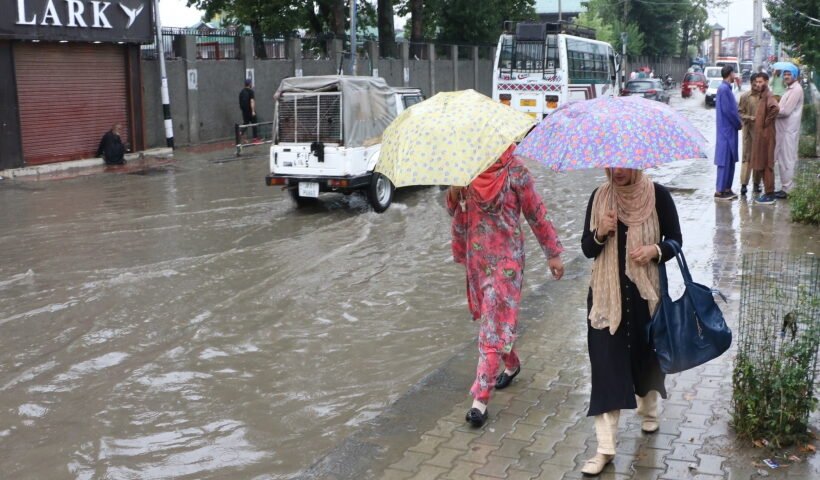 Women shield themselves with umbrellas as they walk past an inundated street during rains